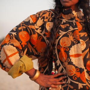 May include: A woman wearing a black embroidered jacket with orange and cream floral designs. She is wearing a gold bangle on her wrist and a beaded necklace.