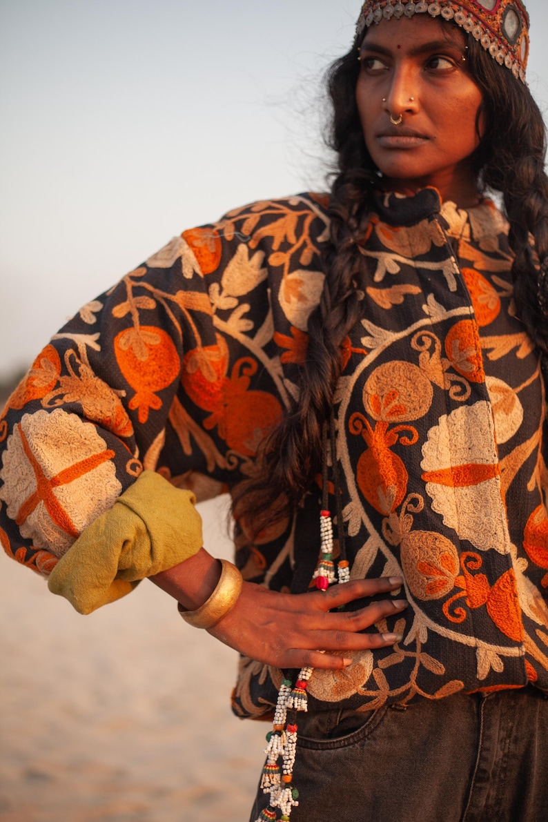 May include: A woman wearing a black embroidered jacket with orange and cream floral designs. She is wearing a gold bangle on her wrist and a beaded necklace.