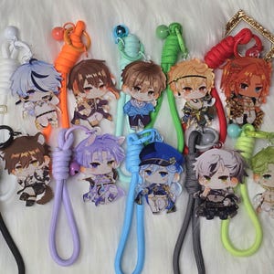 May include: A collection of eleven acrylic character keychains, each attached to a matching colored paracord lanyard. The anime-style figures feature diverse hair colors and detailed outfits, with small charms like scissors and bells attached.