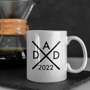 Könnte beinhalten: Weiße Keramiktasse mit einem schwarzen grafischen Design. Das Design zeigt zwei gekreuzte Linien mit den Buchstaben "D" und "A" an den Enden der Linien. Das Jahr "2022" ist unter dem Design gedruckt.