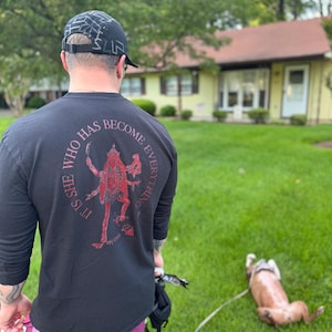 May include: Black long-sleeve shirt with a red graphic and the text "IT'S SHE WHO HAS BECOME EVERYTHING". The person is wearing a black cap and maroon shorts. A dog is lying on the grass.