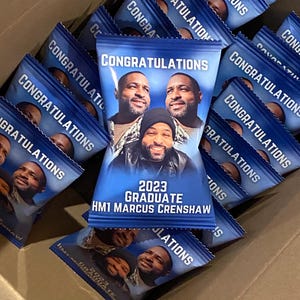 May include: A blue and white bag with the text "Congratulations" and "2023 Graduate HM1 Marcus Crenshaw". The bag has a picture of three men on it.