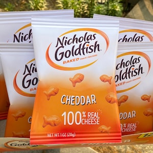 May include: A box of 12 individual packs of  Nicholas Goldfish Cheddar Baked Snack Crackers. Each pack contains 1 ounce (28 grams) of crackers and is labeled "100% Real Cheese".