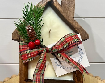 May include: A wooden house-shaped candle with a white candle inside. The candle is decorated with a red and green plaid bow and a sprig of greenery with red berries. The candle is labeled "Riverhouse Candles".