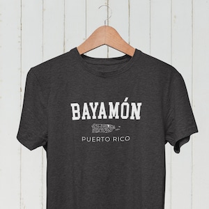 May include: A dark grey t-shirt with white text that reads "BAYAMÓN PUERTO RICO". The text is in a collegiate font. A white outline of the island of Puerto Rico is below the text.