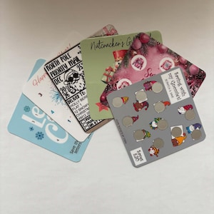 May include: A collection of five rectangular savings challenge cards with various holiday themes. One card features a grey background with gnome illustrations and scratch-off circles, labelled Saving with my Gnomies! Scratch & Save. Another card is a black and white North Pole Priority Mail envelope design. Others include a Nutcracker theme and blue winter snowflake patterns.