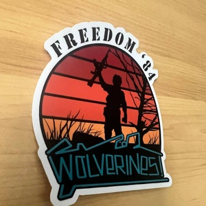 May include: A black and white sticker with a silhouette of a person holding a rifle, standing in front of a sunset with the text "FREEDOM '84" above and "WOLVERINES!" below.