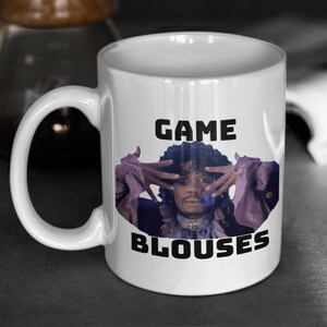 May include: White ceramic mug featuring a portrait of a person with hands up, with the words "GAME BLOUSES" in black block letters. The person in the portrait is wearing a purple ruffled shirt.