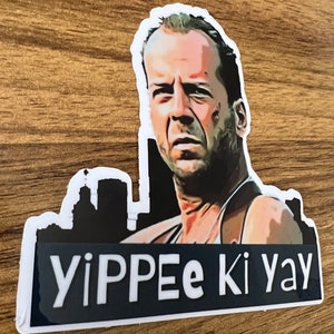 May include: A sticker featuring a black and white silhouette of a city skyline with a man's face in the foreground. The man is wearing a black tank top and has a serious expression. The text "YIPPEE ki yay" is printed in white on a black background.