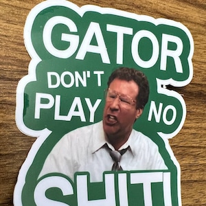 May include: Green and white sticker with a picture of a man with a serious expression. The sticker says "Gator Don't Play No Shit".