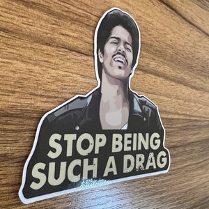 May include: A sticker featuring a cartoon illustration of a man with dark hair and a mustache wearing a black leather jacket. The text "STOP BEING SUCH A DRAG" is printed in gold letters on a black background.