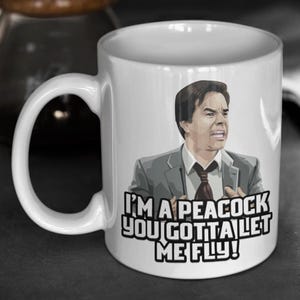 May include: White ceramic mug featuring a cartoon image of a man in a suit with the text "I'M A PEACOCK YOU GOTTA LET ME FLY!". The mug has a large, curved handle and is suitable for hot or cold beverages.