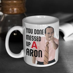 May include: White ceramic mug with the text "YOU DONE MESSED UP A ARON" and an image of a man pointing. The mug has a large handle and is set against a grayscale background.