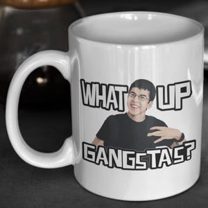 May include: White ceramic mug with a black handle featuring the text "WHAT UP GANGSTAS?" and an illustration of a smiling person wearing glasses and a black shirt. The mug is cylindrical and appears to be 11 ounces.