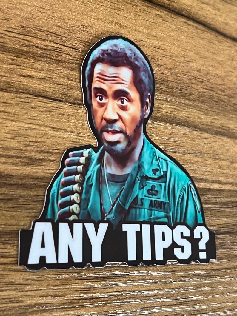 May include: A sticker featuring a man in a green military uniform with the text "ANY TIPS?" below. The man is looking directly at the viewer with a serious expression. He is wearing a necklace and has a bandolier of bullets across his chest. The sticker has a black border.