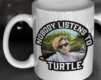 Può includere: Tazza in ceramica bianca con grafica in bianco e nero. La grafica presenta una persona con occhiali da sole in un'auto con la scritta "NOBODY LISTENS TO TURTLE". La tazza ha un manico curvo.