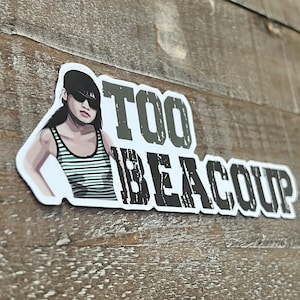 May include: A white sticker with a cartoon illustration of a woman wearing sunglasses and a striped shirt. The text "TOO BEACOUP" is printed in black and white.