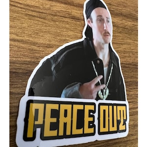 May include: A sticker featuring a man wearing a black jacket and a gold chain, with the text "Peace Out" in yellow letters on a black background.