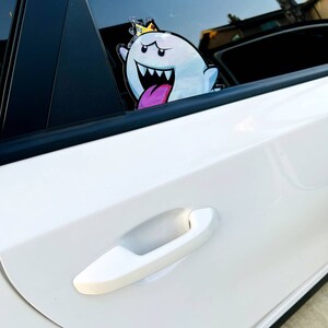 May include: A white car with a cartoon ghost sticker on the back window. The ghost is wearing a gold crown and has a pink tongue sticking out. The car has a white door handle.