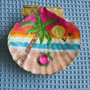 May include: Hand-painted seashell depicting a beach scene. The artwork includes a pink sky, a yellow sun, a green palm tree, and a pink gem. The shell has a gold-painted edge and is set against a blue textured background.