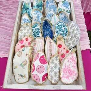 May include: A display of decorative oyster shells in a white wooden tray. The shells are adorned with diverse fabric patterns, such as floral, strawberry, and smiley face motifs. The edges of the shells are finished with gold paint, providing a refined look.