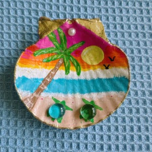 May include: Hand-painted seashell trinket dish depicting a tropical beach scene with a palm tree, sunset, and two glass turtle accents. The shell has a gold-painted edge and a pearl accent.