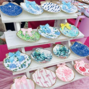 May include: A selection of decorative seashell trinket dishes. Each shell has a unique design, including floral patterns, stripes, and bow accents. Some shells have a pearl in the centre. The shells are arranged on a white tiered shelf, with a pink tablecloth.