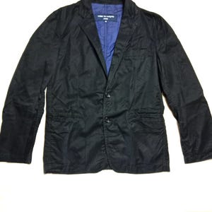 Pode incluir: Um blazer preto com fecho de dois botões e lapelas entalhadas. O forro interior é azul vibrante. O blazer tem dois bolsos frontais e um corte clássico. A etiqueta diz "COMME des GARÇONS HOMME".