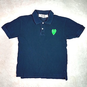 Puede incluir: Polo de color azul marino con un corazón verde en el pecho. Tiene cuello, mangas cortas y tapeta de tres botones. La etiqueta dice "PLAY".