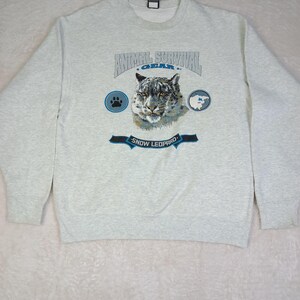以下が含まれることがあります： 雪ヒョウのグラフィックが特徴のライトグレーのスウェットシャツ。「ANIMAL SURVIVAL GEAR」の文字が雪ヒョウの顔の上に、「SNOW LEOPARD」の文字が下に配置されています。足跡と山の風景が描かれた円形のグラフィックも含まれています。