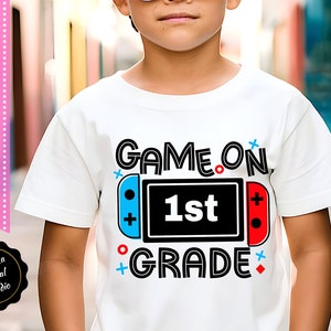 May include: White t-shirt with a black and white graphic design of a video game controller with the text "GAME ON 1st GRADE".