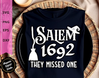 May include: A sleeveless navy blue t-shirt with the text "Salem 1692 They Missed One" in white. The design includes a witch's hat and a broom. The shirt is displayed on a black plate within a woven basket.