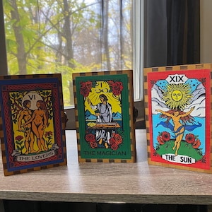 May include: Three tarot cards with colourful illustrations and gold trim. The cards are titled "The Lovers", "The Magician", and "The Sun".