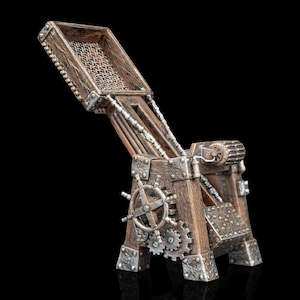 May include: A brown wooden catapult model featuring silver metallic gears, a ship's wheel, and chain details. The structure has a raised throwing arm with a basket, reinforced corners, and a mechanical crank system on the side, all set against a black background.