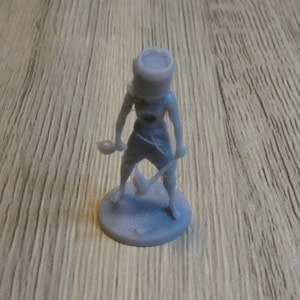 May include: A gray miniature figurine of a person in a military-style outfit, holding a sword and a bowl. The figure is standing on a circular base. The background is a wooden surface.