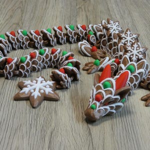 Puede incluir: Un dragón de pan de jengibre marrón con detalles de glaseado blanco, caramelos rojos y verdes y adornos de copo de nieve. El dragón está posado sobre una superficie de madera, mostrando su diseño articulado y su tema festivo.