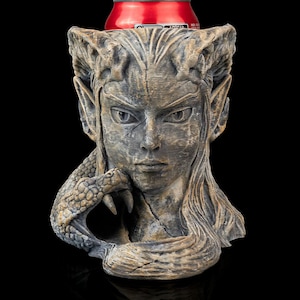 May include: A grey stone sculpture of a woman's head with a dragon's claw holding her chin. The sculpture is a drink holder with a red can inside.