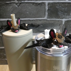 Puede incluir: Dos tapones para bebidas con forma de murciélago con cara marrón, orejas rosas y boca rosa. Las alas del murciélago son negras. Un tapón está sobre un vaso beige, el otro sobre un vaso de acero inoxidable.