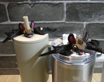 May include: Two bat-shaped drink toppers with a brown face, pink ears, and a pink mouth. The bat wings are black. One topper is on a beige tumbler, the other on a stainless steel tumbler.