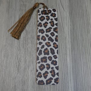 Puede incluir: Marcapáginas rectangular con estampado de leopardo en manchas marrones y negras sobre fondo blanco. Tiene una borla dorada en la parte superior. El marcapáginas está hecho de un material resistente.