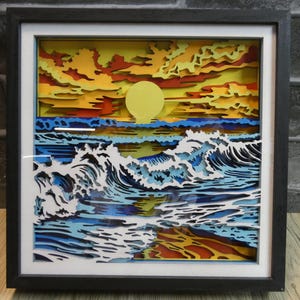 May include: A framed, layered paper art piece depicting a sunset over the ocean. The artwork features a yellow sun, orange and yellow clouds, and blue and white waves. The frame is black.