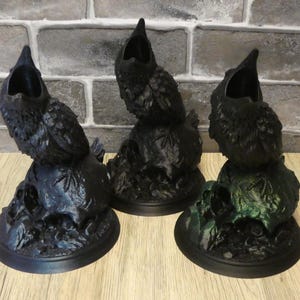 May include: Three black crow figurines with open beaks perched atop skulls. The skulls are on circular bases. The base of the rightmost figurine is painted green.