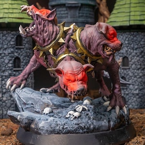 May include: A three-headed, purple-skinned Cerberus statue with red eyes and gold spiked collars. The mythical beast is posed on a rocky base with small skulls scattered around. The background features a castle.