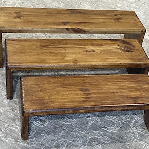 May include: Three wooden display risers in varying sizes. The risers are stained a warm brown colour, showcasing the natural wood grain. Each riser has a rectangular top and four sturdy legs, perfect for showcasing products.