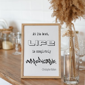May include: A framed print with a black and white quote that reads "At its best, life is completely unpredictable. -Christopher Walken". The print is sitting on a wooden surface with a vase of dried flowers in the background.
