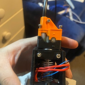 Könnte beinhalten: Ein 3D-gedruckter Extruder für einen 3D-Drucker. Der Extruder ist orange und schwarz und hat eine Feder und ein Zahnrad. Der Extruder ist auf einer weißen Basis montiert.