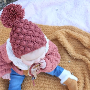 May include: A baby wearing a pink crocheted hat with a pom pom, a pink and white jacket, blue jeans, and white socks. The baby is sitting on a fuzzy blanket.