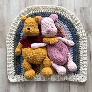 May include: Two crocheted plush toys, a yellow bear with a red shirt and a pink piglet, resting on a semi-circular crocheted rug featuring concentric bands of cream, dark green, blue, and muted orange yarn, all crafted with a textured stitch.