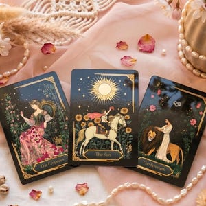 Könnte beinhalten: Drei Tarotkarten mit goldenen Rändern und Illustrationen von Figuren und Symbolen werden auf einem rosa Stoff präsentiert. Die Karten tragen die Aufschriften "The Empress", "The Sun" und "The Strength". Dekorative Elemente sind Blumen, Perlen und Makramee.
