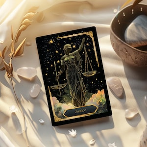 May include: A tarot card featuring the Justice card with a celestial background and a golden-colored illustration of a woman holding scales. The card is set against a backdrop of white fabric, with rose quartz crystals and a decorative bowl.